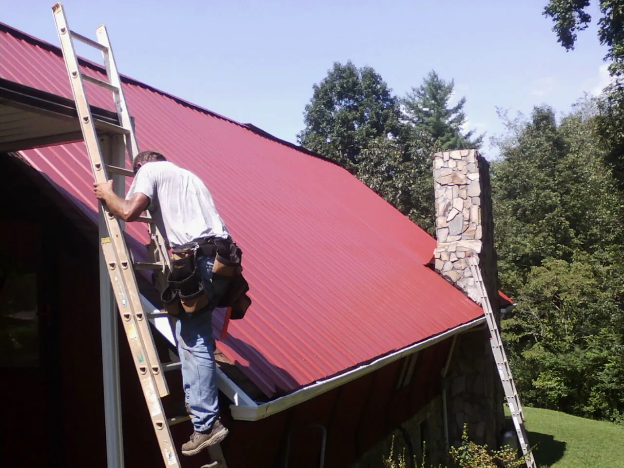 Roofer on a ladder beside a red metal roof and stone chimney — Soffit & Fascia Repair by Roofing Brothers in Thayer
